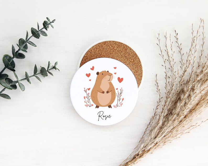 Puede incluir: Posavasos de cer&aacute;mica blanco con un h&aacute;mster de dibujos animados marr&oacute;n y blanco con corazones rojos y el nombre Rosie impreso en la parte inferior.