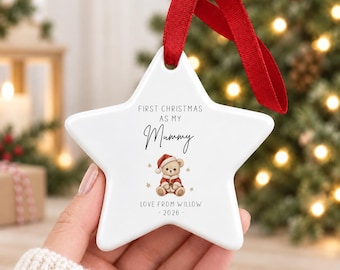 Puede incluir: Un adorno de cerámica blanco en forma de estrella con una cinta roja para colgar. El texto dice FIRST CHRISTMAS AS MY Mummy, LOVE FROM WILLOW y 2026. En el centro hay una ilustración de un osito de peluche con gorro y abrigo de Papá Noel.