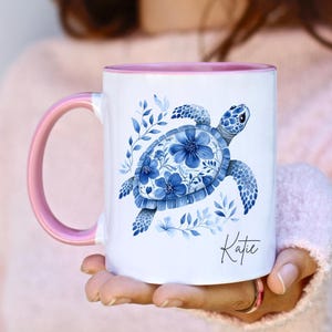 Può includere: Tazza in ceramica bianca con manico e interno rosa. La tazza presenta un disegno di tartaruga marina blu con accenti floreali e il nome "Katie" in corsivo. Il design è su sfondo bianco.