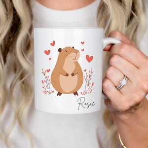 Puede incluir: Taza de cer&aacute;mica blanca con una ilustraci&oacute;n de dibujos animados de un h&aacute;mster marr&oacute;n con corazones rosas y flores. La taza tiene el nombre "Rosie" impreso en la parte inferior.