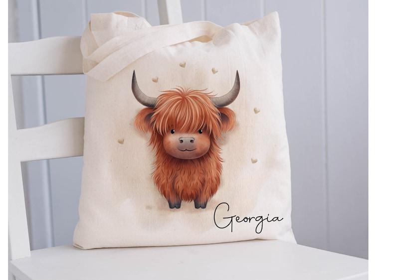 May include: A white canvas tote bag with a brown and white Highland cow illustration and the name "Georgia" printed on the bottom. The bag has a brown handle and is decorated with small gold hearts.