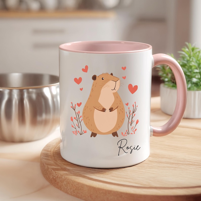 Puede incluir: Taza de cer&aacute;mica blanca con borde y asa de color rosa. La taza presenta una ilustraci&oacute;n de dibujos animados de un h&aacute;mster marr&oacute;n con corazones rosas y flores. El nombre "Rosie" est&aacute; impreso en la taza.