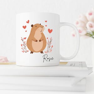 Puede incluir: Taza de cer&aacute;mica blanca con un h&aacute;mster de dibujos animados marr&oacute;n con corazones rosas y el nombre "Rosie" impreso en la taza.