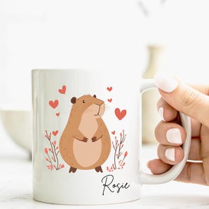 Puede incluir: Taza de cer&aacute;mica blanca con un roedor de dibujos animados marr&oacute;n, corazones rojos y detalles florales rosas. La taza tiene el nombre "Rosie" escrito a mano en negro en la parte inferior.