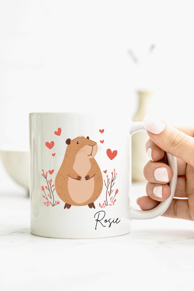 Puede incluir: Taza de cer&aacute;mica blanca con un roedor de dibujos animados marr&oacute;n, corazones rojos y detalles florales rosas. La taza tiene el nombre "Rosie" escrito a mano en negro en la parte inferior.