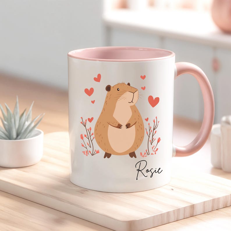 Puede incluir: Una taza de cer&aacute;mica blanca con un borde y un asa de color rosa. La taza presenta una ilustraci&oacute;n de dibujos animados de un conejillo de indias marr&oacute;n con corazones rosas y flores. El nombre "Rosie" est&aacute; escrito en cursiva en la parte inferior de la taza.