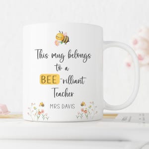 May include: White ceramic mug with a handle, featuring the text "This mug belongs to a BEE-rilliant Teacher" and a cartoon bee illustration. Floral and bee accents are at the bottom. The name "MRS DAVIS" is printed below the text.
