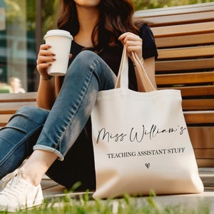 May include: A white canvas tote bag with black text that reads "Miss Williams' Teaching Assistant Stuff" with a small heart symbol below the text. The bag is being held by a person wearing blue jeans and a black shirt.