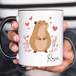 Puede incluir: Taza de cer&aacute;mica blanca con asa negra. La taza presenta una ilustraci&oacute;n de capibara marr&oacute;n de dibujos animados con corazones rojos y el nombre "Rosie" impreso en la parte inferior.