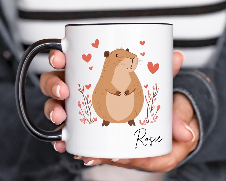 Puede incluir: Taza de cer&aacute;mica blanca con asa negra. La taza presenta una ilustraci&oacute;n de capibara marr&oacute;n de dibujos animados con corazones rojos y el nombre "Rosie" impreso en la parte inferior.