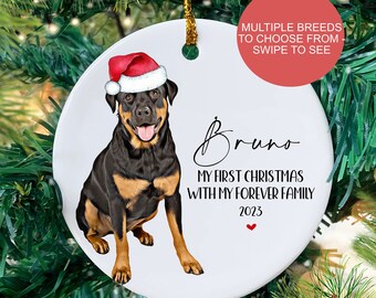 May include: White ceramic Christmas ornament with a black and brown Rottweiler dog wearing a red Santa hat. The ornament has the text "Bruno, My First Christmas with My Forever Family, 2023" on it.