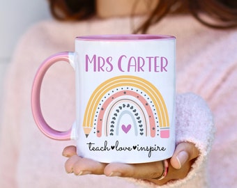 May include: White ceramic mug with a pink rim and handle. The mug features a rainbow design with yellow, pink, and orange stripes, black hearts, and stars. The text "MRS CARTER" is printed in purple above the rainbow. The text "teach love inspire" is printed in black below the rainbow.
