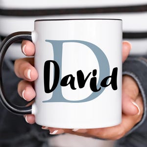 May include: White ceramic mug with a black handle. The mug has a blue letter D and the name "David" written in black cursive.