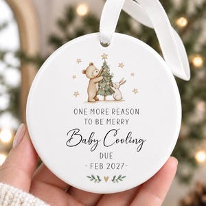 May include: A round white ceramic ornament with a white ribbon hanger. The design features a brown bear and a white rabbit decorating a Christmas tree with stars. Text reads: ONE MORE REASON TO BE MERRY Baby Cooling DUE - FEB 2027 -.
