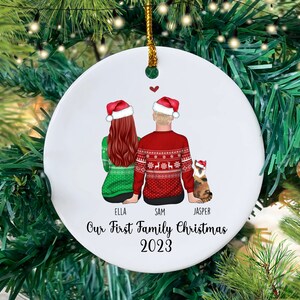 May include: White round ceramic bauble featuring a couple in red and green Christmas jumpers and a cat wearing a Santa hat. The text reads "Our First Family Christmas 2023" with the names Ella, Sam, and Jasper.