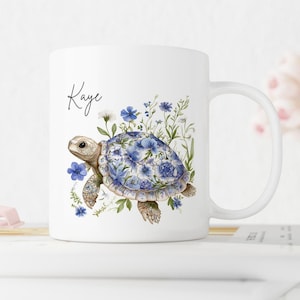 Può includere: Tazza in ceramica bianca con un'illustrazione ad acquerello di una tartaruga con un motivo floreale blu. La tazza ha un manico e il nome "Kaye" scritto in corsivo.