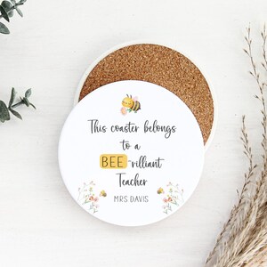 May include: A round white coaster with a cork base. The coaster features the text "This coaster belongs to a BEE-rilliant Teacher" with a bee graphic and floral accents. The name "MRS DAVIS" is printed below.