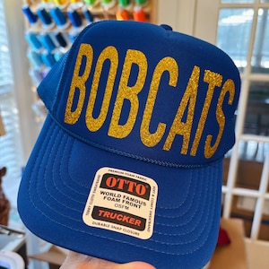 May include: A blue trucker hat with the word "BOBCATS" in gold glitter lettering. The hat has a mesh back and a snap closure. The front of the hat has a label that says "OTTO WORLD FAMOUS FOAM FRONT".
