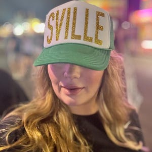 May include: A woman wearing a green and white trucker hat with gold glitter lettering that says "SVILLE".
