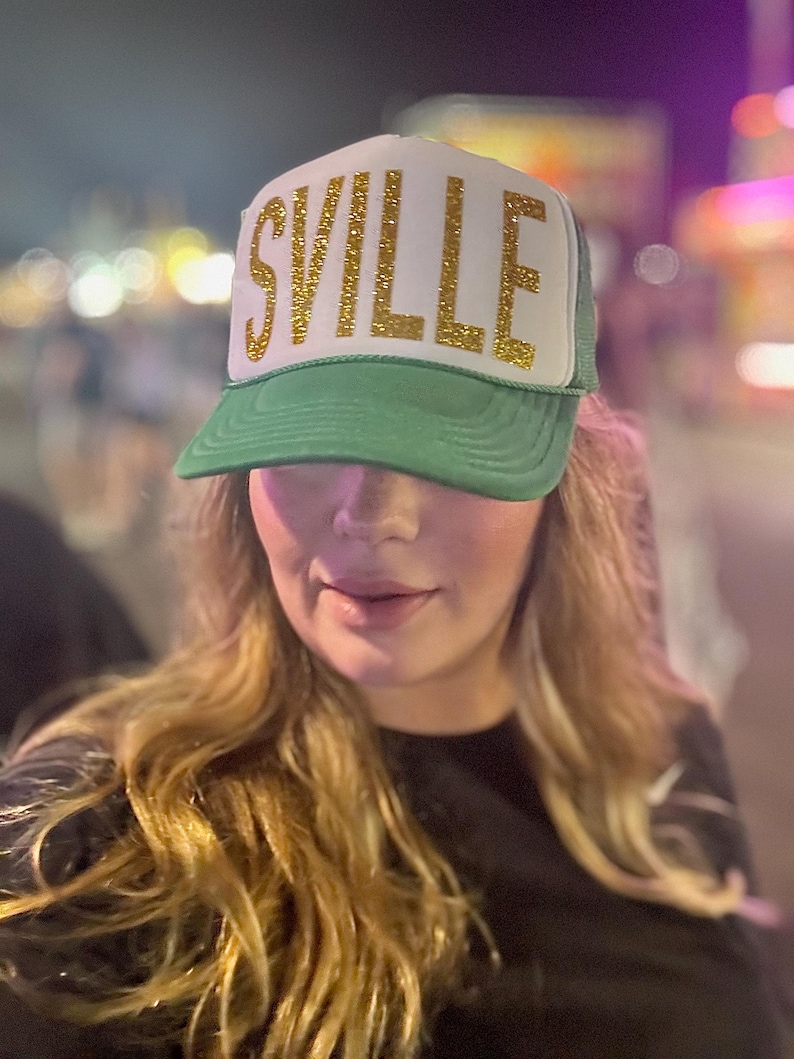 May include: A woman wearing a green and white trucker hat with gold glitter lettering that says "SVILLE".