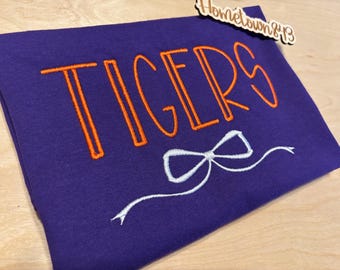 May include: A purple textile item with the word "TIGERS" embroidered in orange, outlined in a lighter orange. A white bow and trailing ribbon are embroidered below the word. A wooden tag with the text "Hometown&13" is attached.