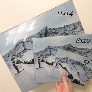 May include: A collection of art prints featuring a snowy mountain landscape with skiers. The prints are in various sizes, including 11x14, 8x10, and 5x7 inches. The artwork depicts a winter scene with a skier in a red jacket.