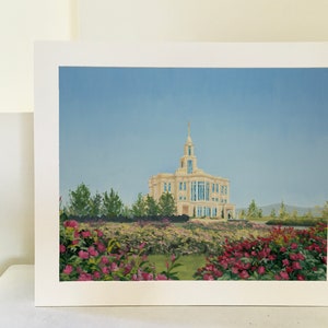 May include: An oil painting of a light-colored building with a tall spire, set against a blue sky. The building is surrounded by green trees and a vibrant garden of pink and red flowers. The artwork is framed in white.