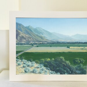 May include: A framed painting depicting a landscape with a valley, mountains in the distance, and a road winding through the valley. The painting is done in a realistic style with muted colors.
