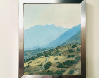 May include: A framed oil painting depicting a landscape with a winding road leading through a valley with rolling hills and distant mountains. The sky is a light blue with a hazy effect. The painting is framed in a silver frame.