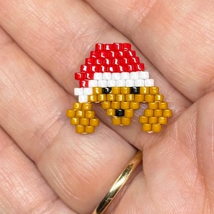 May include: A small, beaded charm in the shape of a bear wearing a Santa hat. The hat is red and white, and the bear is yellow with black eyes. The charm is made of small, colorful beads.