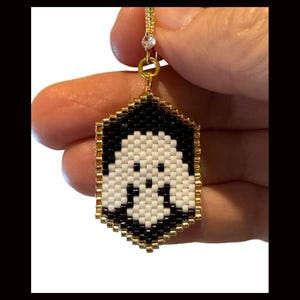 May include: A black and white beaded ghost charm with gold accents. The charm is hanging from a gold chain with a clear crystal bead.