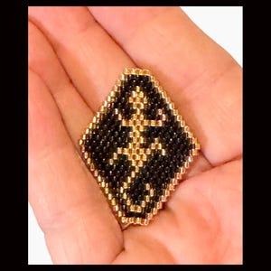 Puede incluir: Un colgante de cuentas negras y doradas en forma de diamante. El colgante presenta un diseño geométrico con un centro dorado y bordes negros.