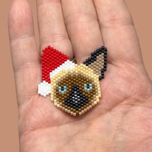 May include: A beaded Siamese cat ornament wearing a red and white Santa hat. The cat's face is tan, black, and white with blue eyes. The ornament is held in a hand.