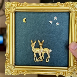 May include: A square gold-toned ornate frame containing a dark teal background. Mounted on the background are two gold-toned deer figures with small white pearl accents, a gold crescent moon, and three small white star-shaped embellishments.