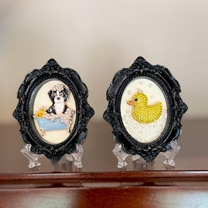 Può includere: Due cornici ovali nere decorate su supporti trasparenti. La cornice sinistra contiene un cane bianco e nero in una vasca da bagno azzurra con schiuma bianca. La cornice destra presenta una papera gialla circondata da piccole perle bianche.