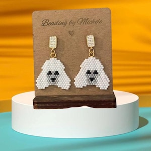 May include: A pair of white beaded earrings with a dog face design. The earrings are hanging from gold hooks with clear crystal accents. The card behind the earrings says "Beading by Michele".