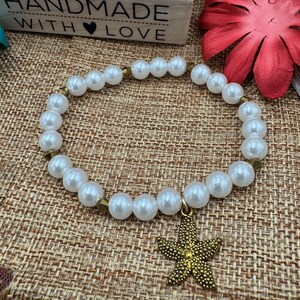Pode incluir: Pulseira de p&eacute;rolas brancas com um berloque de estrela do mar dourada. A pulseira apresenta p&eacute;rolas brancas grandes e redondas unidas por espa&ccedil;adores dourados. O berloque de estrela do mar tem uma superf&iacute;cie texturizada.