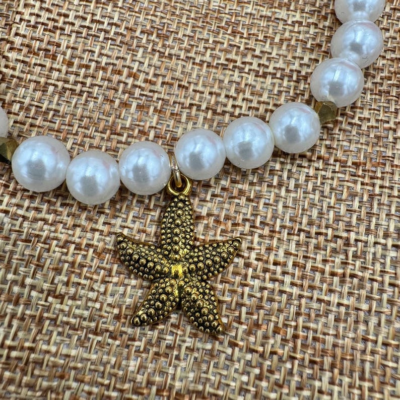 Pode incluir: Um colar de p&eacute;rolas com um pingente de estrela do mar dourada. O colar apresenta p&eacute;rolas brancas grandes e redondas. O pingente de estrela do mar &eacute; texturizado e tem um acabamento dourado. O colar &eacute; exibido em uma superf&iacute;cie tecida bege.