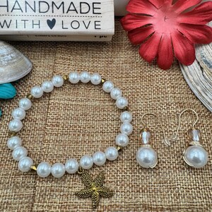 Pode incluir: Uma pulseira de p&eacute;rolas com um pingente de estrela do mar dourada e brincos de p&eacute;rolas combinando. A pulseira e os brincos t&ecirc;m p&eacute;rolas brancas e detalhes dourados. A imagem tamb&eacute;m inclui um carimbo de madeira que diz "HANDMADE WITH LOVE".