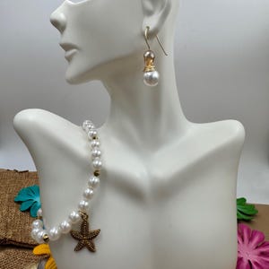 Pode incluir: Um conjunto de colar e brincos de p&eacute;rolas exibido em um manequim branco. O colar apresenta p&eacute;rolas brancas com detalhes dourados e um pingente de estrela do mar de bronze. Os brincos t&ecirc;m um gancho dourado com uma gota de p&eacute;rola. O conjunto de joias &eacute; projetado para um tema de praia ou n&aacute;utico.
