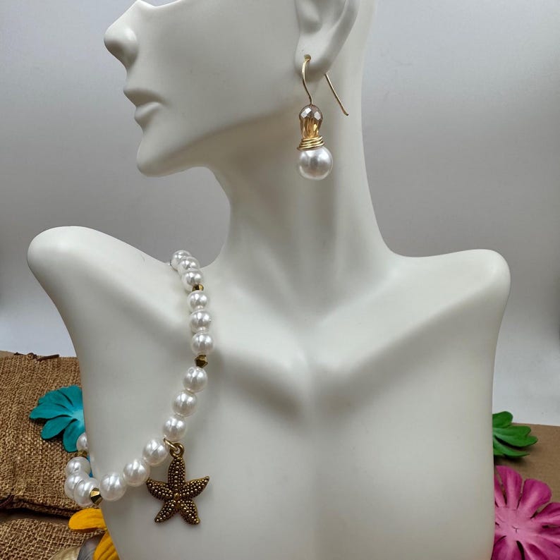 Pode incluir: Um conjunto de colar e brincos de p&eacute;rolas exibido em um manequim branco. O colar apresenta p&eacute;rolas brancas com detalhes dourados e um pingente de estrela do mar de bronze. Os brincos t&ecirc;m um gancho dourado com uma gota de p&eacute;rola. O conjunto de joias &eacute; projetado para um tema de praia ou n&aacute;utico.
