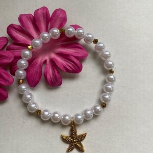 Pode incluir: Uma pulseira de p&eacute;rolas brancas com detalhes dourados e um pingente de estrela do mar dourada. A pulseira est&aacute; sobre uma flor rosa. As p&eacute;rolas s&atilde;o redondas e espa&ccedil;adas uniformemente. O pingente de estrela do mar &eacute; detalhado e texturizado.