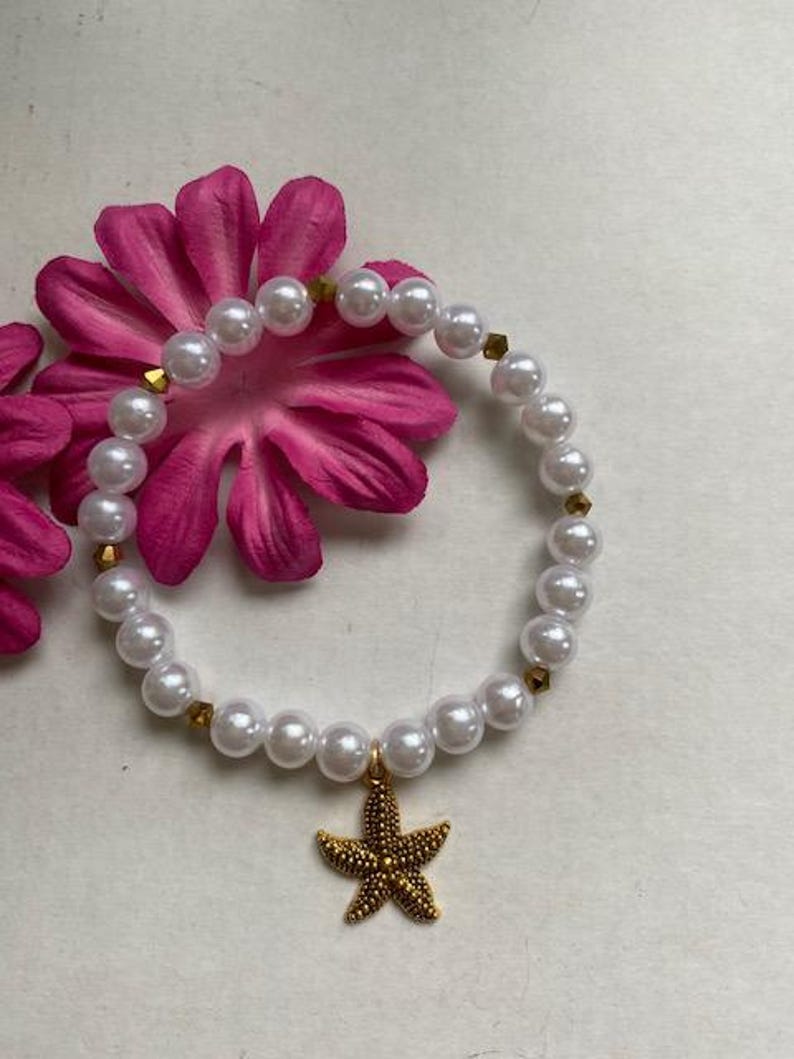 Pode incluir: Uma pulseira de p&eacute;rolas brancas com detalhes dourados e um pingente de estrela do mar dourada. A pulseira est&aacute; sobre uma flor rosa. As p&eacute;rolas s&atilde;o redondas e espa&ccedil;adas uniformemente. O pingente de estrela do mar &eacute; detalhado e texturizado.