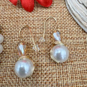 Pode incluir: Um par de brincos pendurados dourados. Cada brinco apresenta uma grande conta branca perolada, encimada por um cristal &acirc;mbar facetado. As contas s&atilde;o enroladas em arame dourado e os brincos t&ecirc;m um fecho de gancho simples.