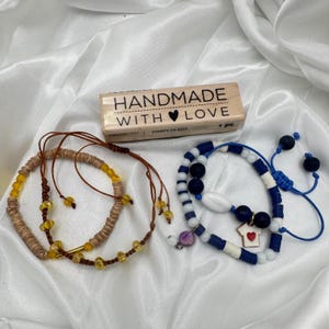 May include: Two handmade bracelets with adjustable cords and a wooden stamp. One bracelet is brown with yellow beads, the other is blue and white with dark blue beads and a small charm with a red heart. The stamp reads "HANDMADE WITH LOVE".