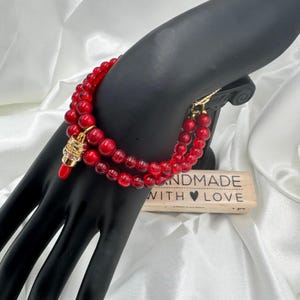 May include: A double-strand red beaded bracelet with a gold-colored charm and clasp. The charm has red and gold accents. The bracelet is displayed on a black mannequin hand. A wooden block with the text "HANDMADE WITH LOVE" is also visible.