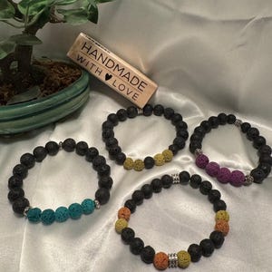 Puede incluir: Cuatro pulseras hechas a mano con piedras de lava y cuentas de acento en turquesa, amarillo, morado y naranja. Cada pulsera presenta cuentas de lava negras y espaciadores plateados. También es visible un sello de madera con el texto "HANDMADE WITH LOVE".