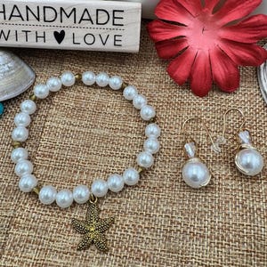Pode incluir: Uma pulseira de p&eacute;rolas feita &agrave; m&atilde;o com um pingente de estrela do mar dourada e brincos de p&eacute;rolas combinando. A pulseira e os brincos s&atilde;o exibidos com um bloco de madeira que diz "HANDMADE WITH LOVE". Uma flor vermelha est&aacute; no fundo.
