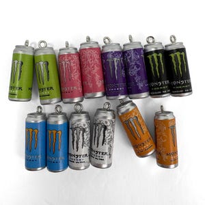 May include: A collection of fourteen miniature energy drink can charms, each featuring a metal loop at the top. The charms display various colors including green, pink, purple, black, blue, white, and orange, all with the Monster Energy logo.