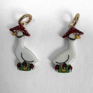 May include: Two white goose charms with yellow beaks, each wearing a red mushroom cap with white spots. The geese are standing on a green patch with small red mushrooms. Each charm features a gold-toned metal jump ring at the top.